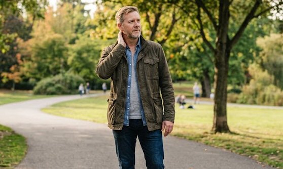 An adult standing on a park path in daylight with one hand resting lightly at the side of the neck after a walk